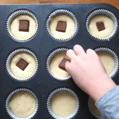 Dziecko dodaje kawałki czekolady do ciasta na muffinki waniliowe. W foremce widać już kilka gotowych do pieczenia babeczek.