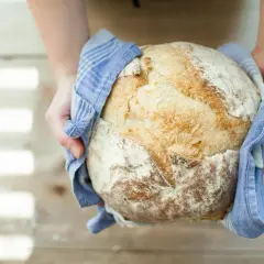 Ręce trzymają świeżo upieczony bochenek chleba, owinięty w niebieski ręcznik. Czy można kroić chleb zaraz po upieczeniu? Lepiej poczekać, aż ostygnie.