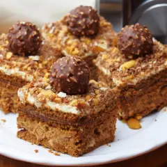 Pyszne ciasto Ferrero Rocher, udekorowane całymi czekoladkami i posypką orzechową, prezentuje się apetycznie na białym talerzu.