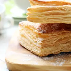 Stos puszystego ciasta francuskiego, idealnego do nauki jak zrobić ciasto francuskie. Posypane cukrem pudrem, na drewnianej desce.