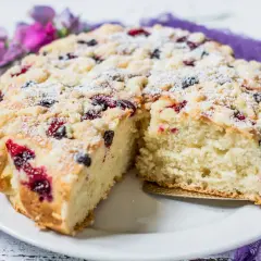 Puszyste ciasto z czarną porzeczką, posypane cukrem pudrem i kruszonką, na białym talerzu, ozdobione fioletowymi kwiatami.