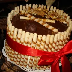 Wyjątkowy tort z rurkami waflowymi, polany czekoladą, ozdobiony orzechami i czerwoną wstążką.