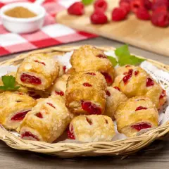 Koszyk pełen chrupiących ciastek z marmoladą z ciasta francuskiego, posypanych cukrem. Obok maliny i dżem.