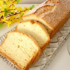 Puszysta babka z białek z olejem, oprószona cukrem pudrem, na szklanej paterze, obok gałązka żółtych kwiatów.