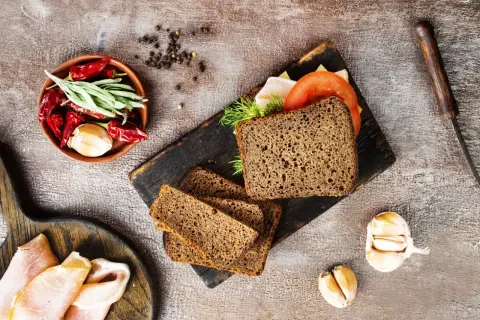 Pyszny chleb z razowej mąki pszennej, idealny na kanapki. Obok składniki: pomidor, ser, wędlina, czosnek, papryczki chili i zioła.