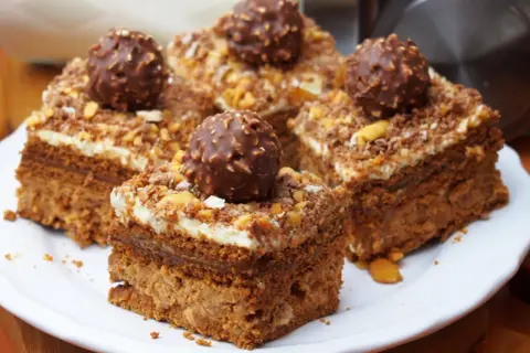 Pyszne ciasto Ferrero Rocher, udekorowane całymi czekoladkami i posypką orzechową, prezentuje się apetycznie na białym talerzu.
