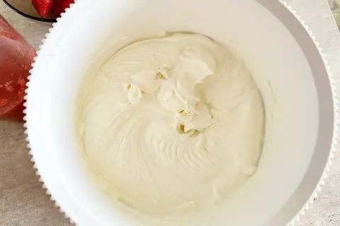 Gładki, puszysty krem z serka mascarpone w białej misie, gotowy do deseru. Obok miski widać świeże truskawki.