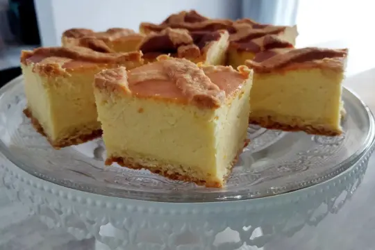 Pyszny sernik na ciasteczkowym spodzie, pokrojony na kwadraty, prezentuje się apetycznie na szklanej paterze.
