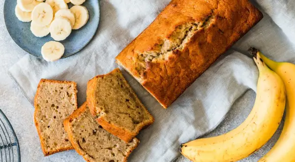 Pyszne ciasto z bananem, pokrojone na kromki, obok świeże banany i talerz z plasterkami.