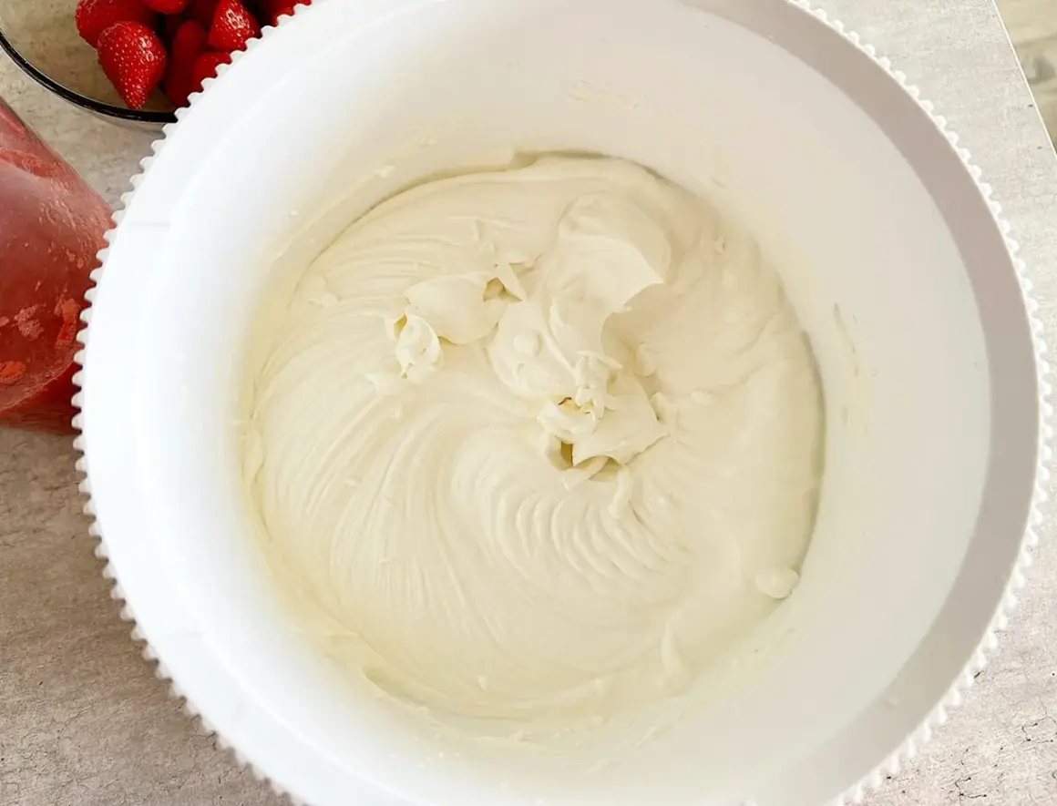 Gładki, puszysty krem z serka mascarpone w białej misie, gotowy do deseru. Obok miski widać świeże truskawki.