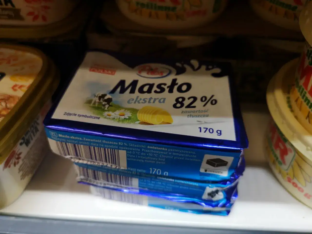Kostka masła ekstra 82% waży 170 g. Na opakowaniu widnieje krowa na łące.
