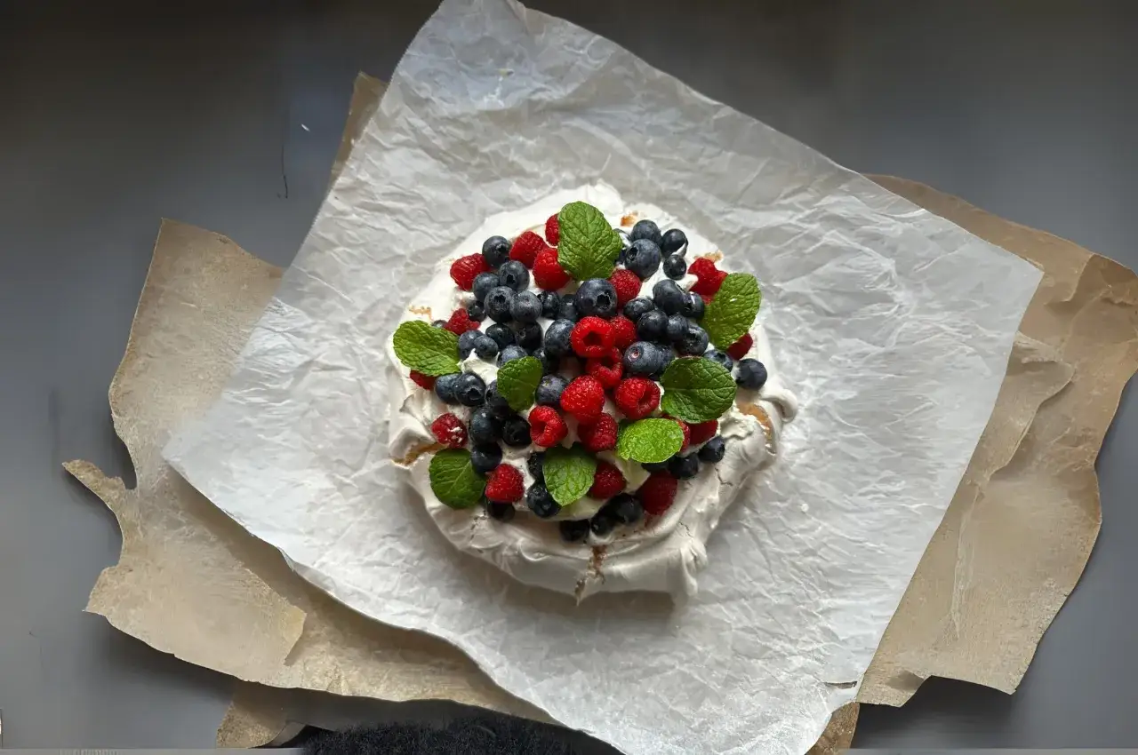 Beza Pavlova z mascarpone, udekorowana malinami, borówkami i miętą, prezentuje się apetycznie na papierze do pieczenia.