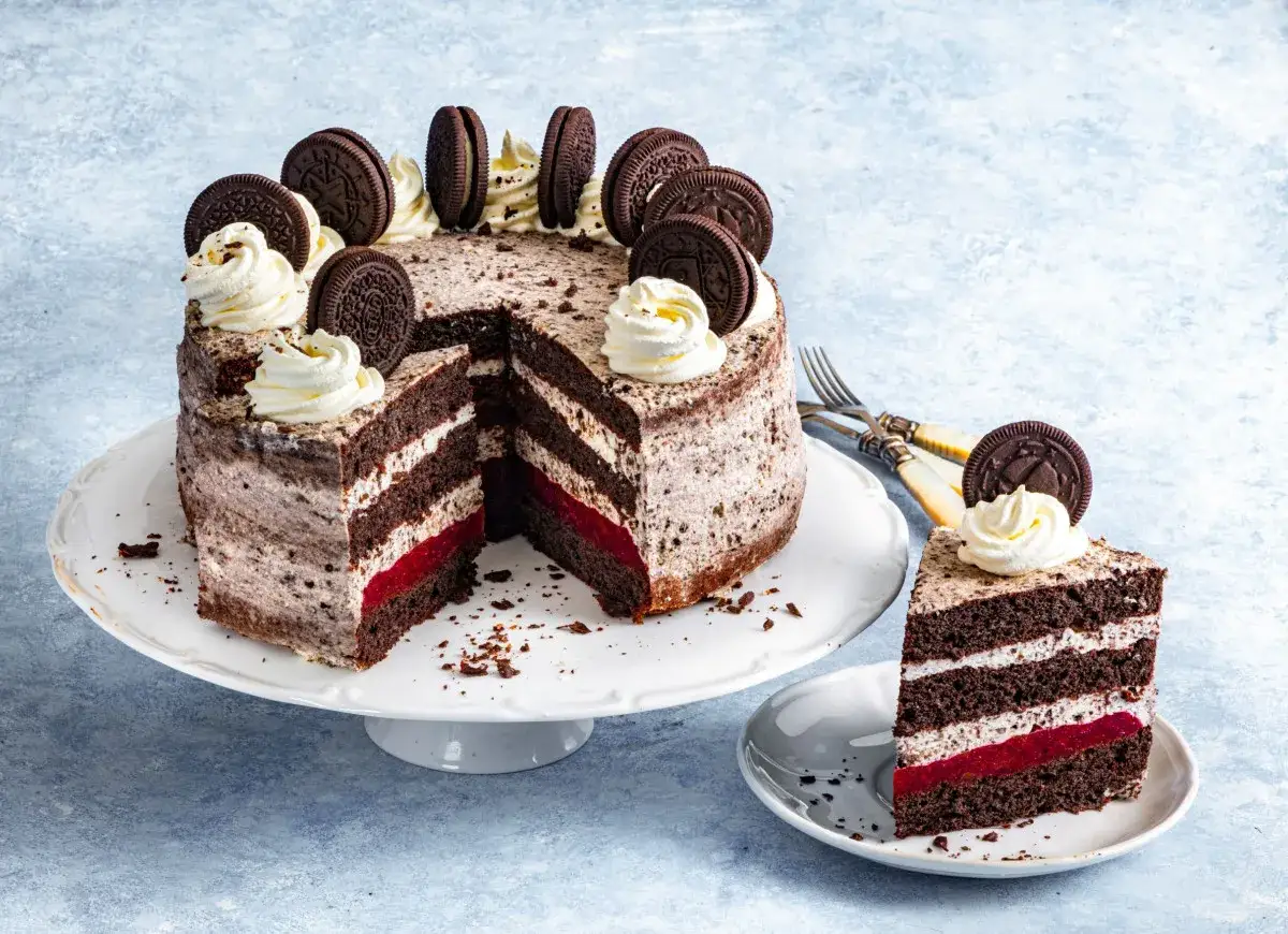 Pyszny tort bez pieczenia z warstwami czekoladowego ciasta, kremu i czerwonego dżemu, ozdobiony ciasteczkami Oreo i bitą śmietaną.