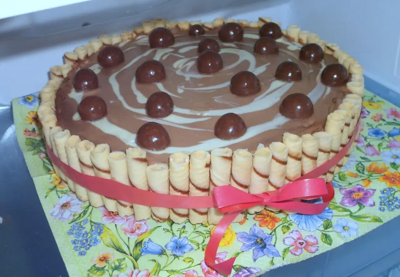 Pyszny tort z rurkami waflowymi i czekoladowymi kulkami, ozdobiony różową wstążką.
