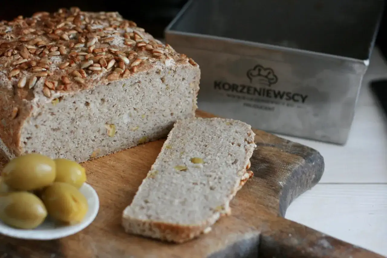 Chleb gryczany z pestkami słonecznika, pokrojony bochenek i kromka na desce, obok oliwki. Czy chleb gryczany jest lekkostrawny? Tak, ten wygląda na taki!