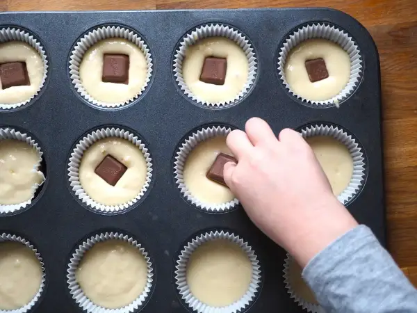Dziecko dodaje kawałki czekolady do ciasta na muffinki waniliowe. W foremce widać już kilka gotowych do pieczenia babeczek.