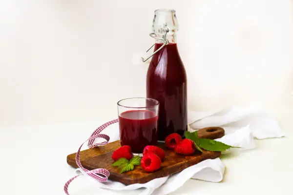 Szklanka i butelka z domowym sokiem z malin, świeże maliny i listki mięty na desce. Idealne na lato, pokazują, jak zrobić sok z malin.