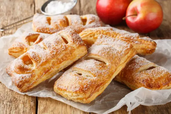 Szybkie ciasto francuskie z jabłkami, posypane cukrem pudrem, leży na papierze. Obok dwa czerwone jabłka.